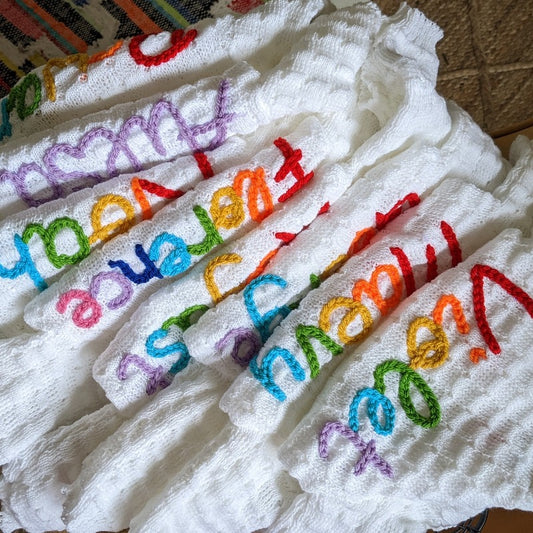 Pile of white knitted baby cardigans with names embroidered in colourful threads, piled on a wooden surface.