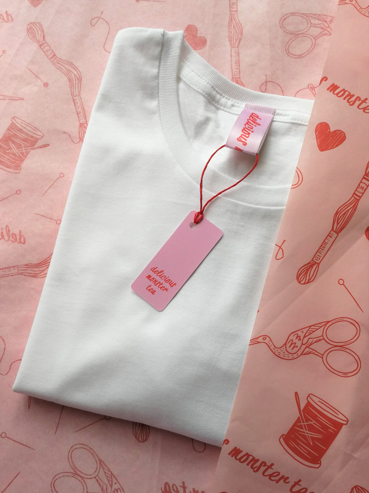 White t-shirt with a pink tag on a pink fabric background with sewing-related illustrations