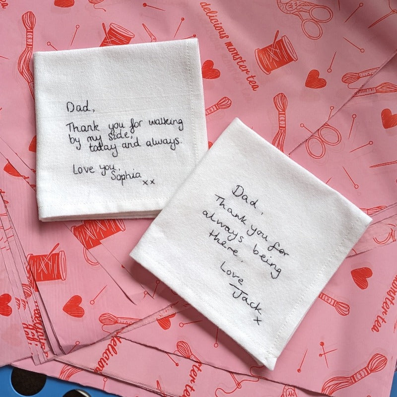 A pair of white cotton handkerchiefs with black embroidered handwritten messages to the father of the bride and father of the groom.