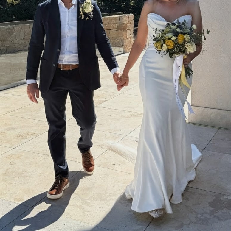 A bride and a groom are walking and holding hands. The bride is holding a bouquet of yellow flowers that are tied with a white satin ribbon. There is a handwritten love note embroidered on the ribbon in blue thread.