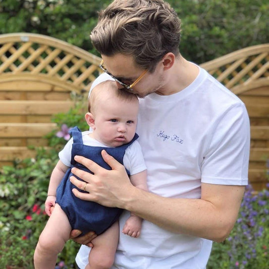 A white man is holding a baby and wearing a white cotton t-shirt that is embroidered with the child's name.