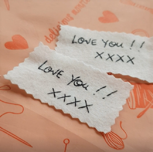 A pair of white cotton wedding dress labels with black embroidered handwriting that reads 'Love you!! xxxx'